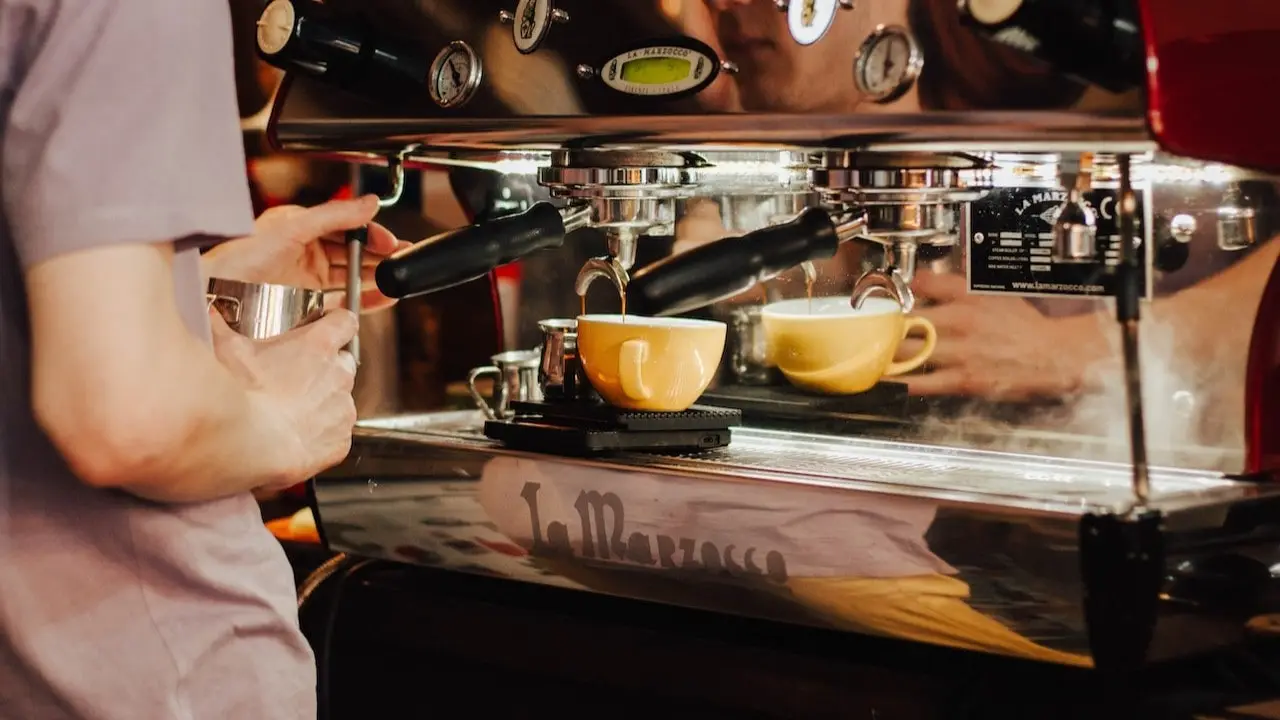 a barista using the milk frother while a coffee machine brews the coffee into a yellow mug